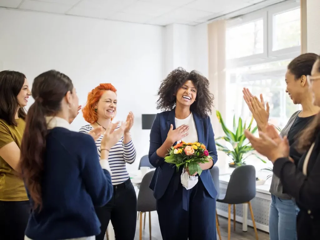 Black woman holding a bouquet of flowers surrounded by coworkers who are clapping for her